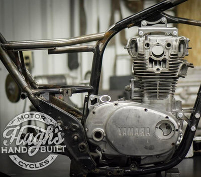 A picture of a motorcycle in a workshop, featuring the Hughs HandBuilt XS650 Top Motor Mount Window 1974-83 for the XS650, designed by Hughs Handbuilt.