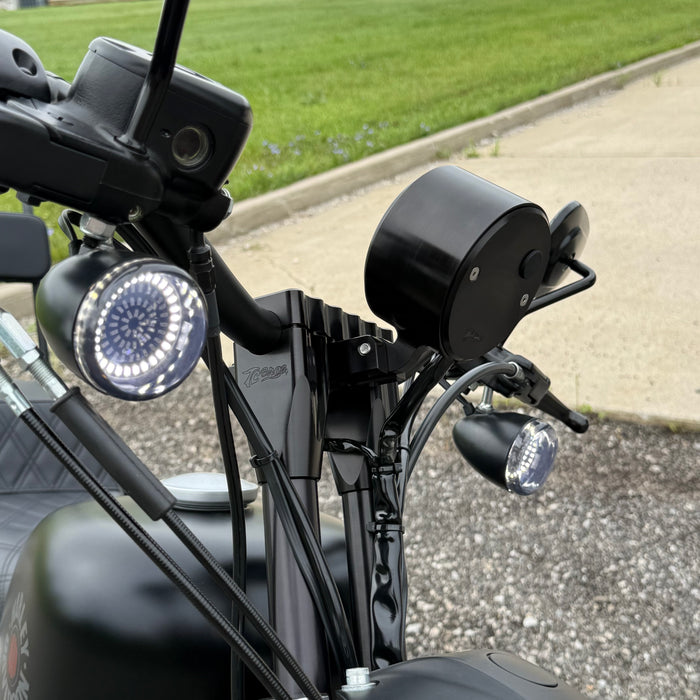 Two cylindrical motorcycle tachometer bezels from TC Bros., including the Pro Series 4 inch Gauge Relocation Bezel for Harley Davidson, in black and silver finishes with similar gauge faces featuring numbers and a red needle.
