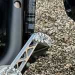 Close-up of a CNC-machined TC Bros. Pro Series MX Shifter/Brake Peg and foot peg assembly, mounted near the engine and exhaust pipes on a concrete surface.