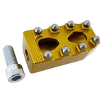 Close-up of a CNC-machined TC Bros. Pro Series MX Shifter/Brake Peg and foot peg assembly, mounted near the engine and exhaust pipes on a concrete surface.