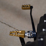 Three TC Bros. Pro Series MX Lite Foot Pegs for Harley Davidson models, featuring metallic spiked surfaces, are arranged side by side in silver, black, and gold.