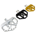 Three TC Bros. Pro Series MX Mini Floorboards for Harley Davidson motorcycles, in silver, black, and gold, are lined up. They resemble bicycle pedals with flat platforms and metal pins for grip.
