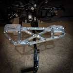 Close-up of TC Bros. Pro Series MX Slim Floorboards for 1984-Up Harley-Davidson Touring, featuring CNC-machined billet aluminum and visible bolts, mounted on a black Harley-Davidson motorcycle on a concrete floor.