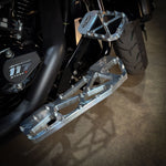 Close-up of TC Bros. Pro Series MX Slim Floorboards for 1984-Up Harley-Davidson Touring, featuring CNC-machined billet aluminum and visible bolts, mounted on a black Harley-Davidson motorcycle on a concrete floor.