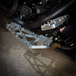 Close-up of TC Bros. Pro Series MX Slim Floorboards for 1984-Up Harley-Davidson Touring, featuring CNC-machined billet aluminum and visible bolts, mounted on a black Harley-Davidson motorcycle on a concrete floor.