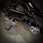 Close-up of TC Bros. Pro Series MX Slim Floorboards for 1984-Up Harley-Davidson Touring, featuring CNC-machined billet aluminum and visible bolts, mounted on a black Harley-Davidson motorcycle on a concrete floor.