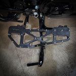 Close-up of TC Bros. Pro Series MX Slim Floorboards for 1984-Up Harley-Davidson Touring, featuring CNC-machined billet aluminum and visible bolts, mounted on a black Harley-Davidson motorcycle on a concrete floor.