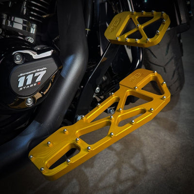 TC Bros. Pro Series MX Slim Floorboards for Harley Davidson Bagger Touring Models feature a skeletal design with metal pins, resembling high-performance pedals in silver, black, and gold. Arranged diagonally, they offer a striking visual appeal.