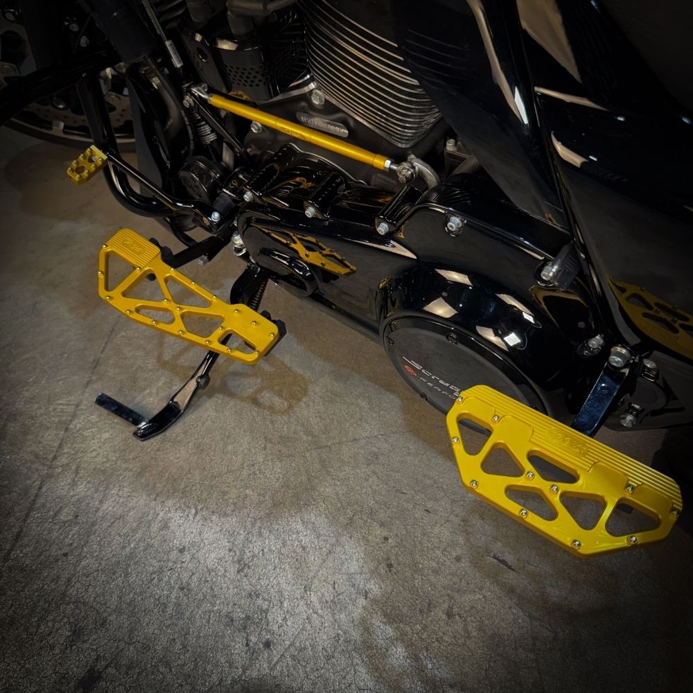 TC Bros. Pro Series MX Slim Floorboards for Harley Davidson Bagger Touring Models feature a skeletal design with metal pins, resembling high-performance pedals in silver, black, and gold. Arranged diagonally, they offer a striking visual appeal.