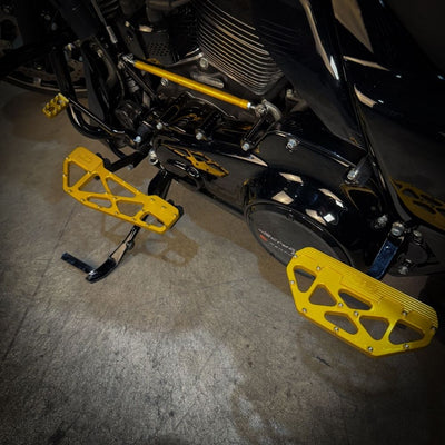 TC Bros. Pro Series MX Slim Floorboards for Harley Davidson Bagger Touring Models feature a skeletal design with metal pins, resembling high-performance pedals in silver, black, and gold. Arranged diagonally, they offer a striking visual appeal.