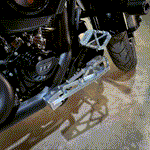 Close-up of a motorcycle's engine and TC Bros. Pro Series Lift Kit for MX Floorboards, with part of the rear wheel on concrete, showcasing improved lean angle.
