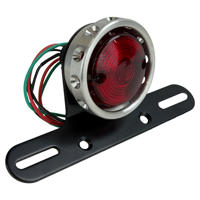 The Mid-USA Vintage Drilled Tail Light by Mid-USA features a round red lens in an aluminum housing, mounted on a black bracket with exposed electrical wires at the back.