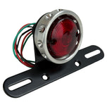 The Mid-USA Vintage Drilled Tail Light by Mid-USA features a round red lens in an aluminum housing, mounted on a black bracket with exposed electrical wires at the back.