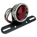 The Mid-USA Vintage Drilled Tail Light by Mid-USA features a round red lens in an aluminum housing, mounted on a black bracket with exposed electrical wires at the back.