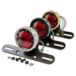 The Mid-USA Vintage Drilled Tail Light by Mid-USA features a round red lens in an aluminum housing, mounted on a black bracket with exposed electrical wires at the back.