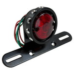 The Mid-USA Vintage Drilled Tail Light by Mid-USA features a round red lens in an aluminum housing, mounted on a black bracket with exposed electrical wires at the back.