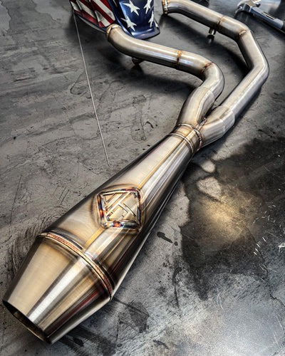 An SP Concepts Lane Splitter Exhaust M8 Softail ST & Sport Glide (TC Bros. Mids) 2018-Present sits on a workshop surface with an American flag design visible in the background.