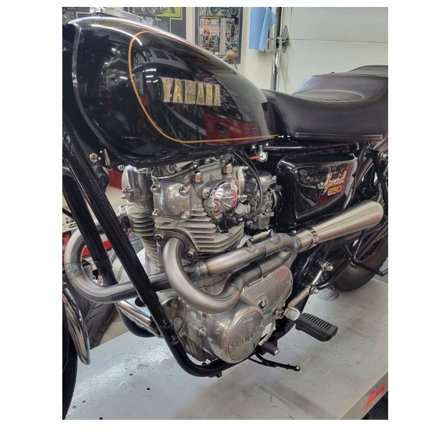 A side view of a Yamaha XS650 motorcycle's Pandemonium 2 into 1 XS650 exhaust system in a workshop setting.