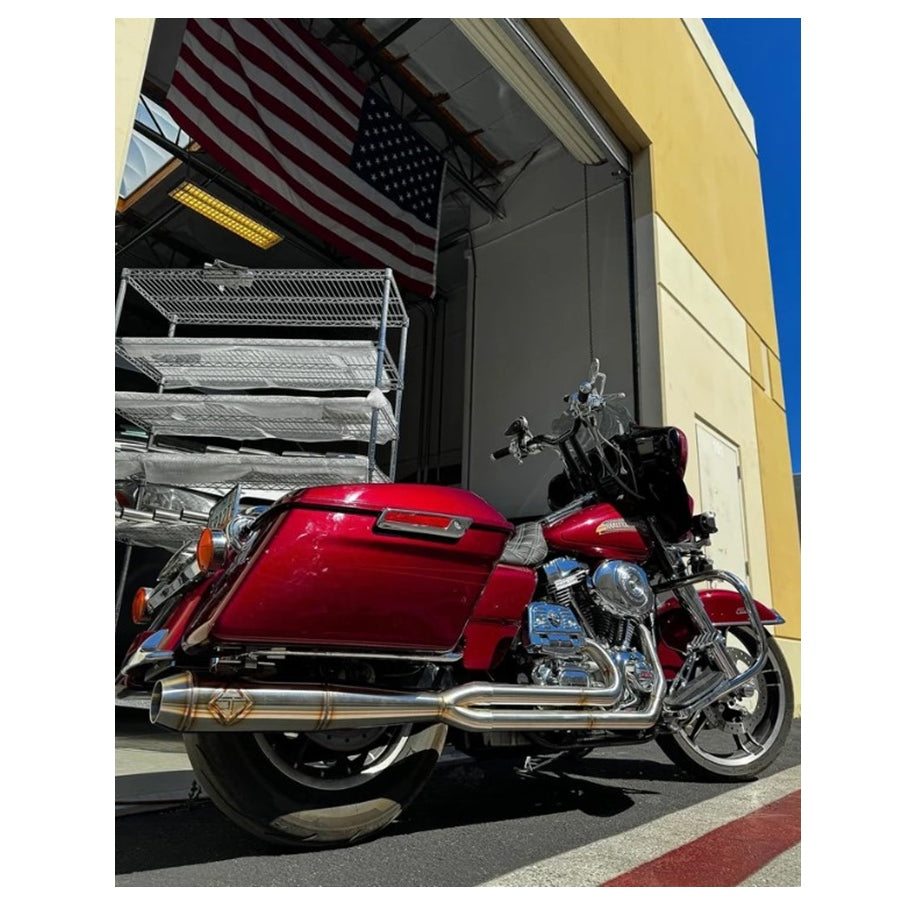 An SP Concepts TC Bagger Full Length Turnpike Exhaust-equipped red motorcycle is parked outside an industrial building with a large American flag hanging inside.