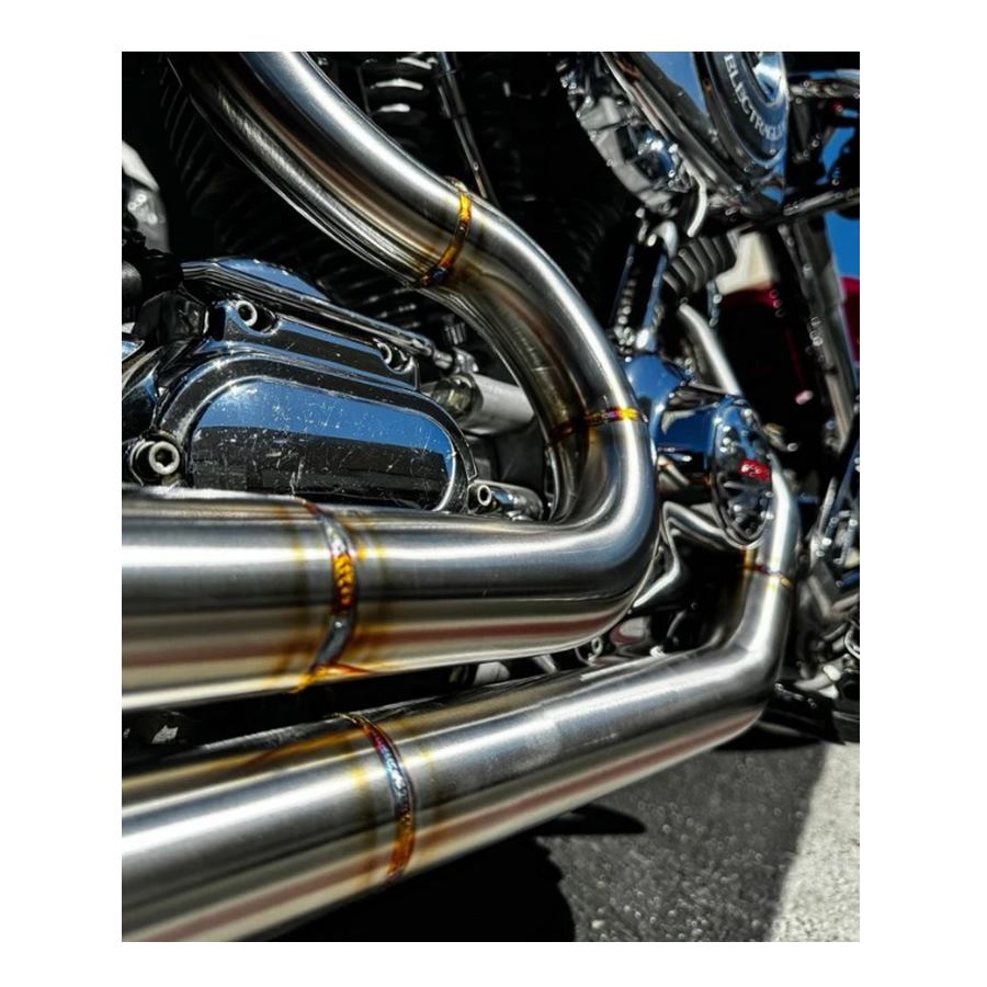 A red motorcycle parked outside a garage, equipped with SP Concepts' gleaming stainless steel TC Bagger Full Length Turnpike Exhaust for 1995-2016 Touring models, sits next to metal racks. Inside, an American flag hangs proudly amidst tools and parts for these touring models.