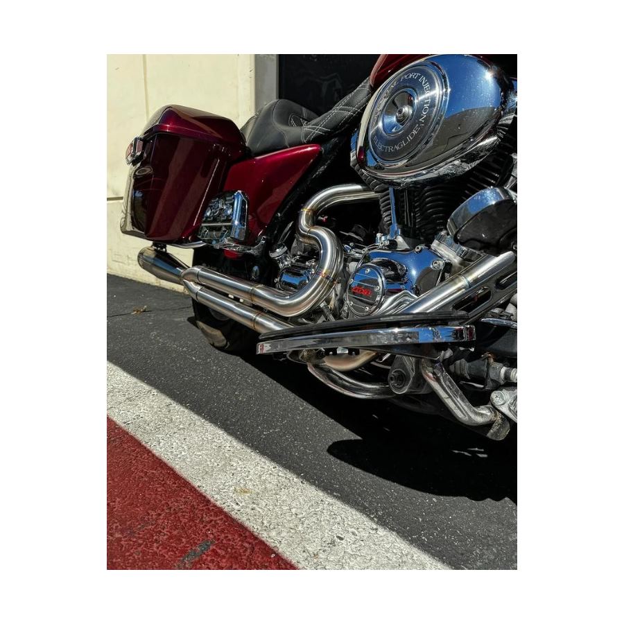 A red motorcycle parked outside a garage, equipped with SP Concepts' gleaming stainless steel TC Bagger Full Length Turnpike Exhaust for 1995-2016 Touring models, sits next to metal racks. Inside, an American flag hangs proudly amidst tools and parts for these touring models.