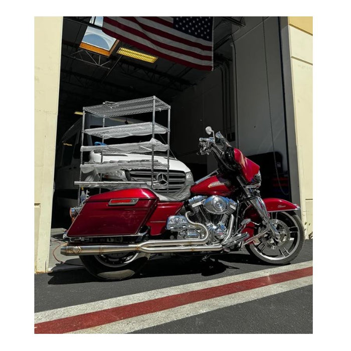 Metal pipes and a Y-shaped exhaust component rest on the concrete floor in a workshop environment, surrounded by shelves and boxes—a scene where SP Concepts works to perfect their Full Length Turnpike Works Edition for Harley &