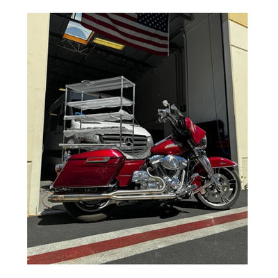 Metal pipes and a Y-shaped exhaust component rest on the concrete floor in a workshop environment, surrounded by shelves and boxes—a scene where SP Concepts works to perfect their Full Length Turnpike Works Edition for Harley '96-'16 Touring exhaust systems.