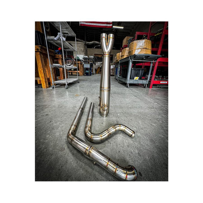 Metal pipes and a Y-shaped exhaust component rest on the concrete floor in a workshop environment, surrounded by shelves and boxes—a scene where SP Concepts works to perfect their Full Length Turnpike Works Edition for Harley &