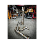 Metal pipes and a Y-shaped exhaust component rest on the concrete floor in a workshop environment, surrounded by shelves and boxes—a scene where SP Concepts works to perfect their Full Length Turnpike Works Edition for Harley &