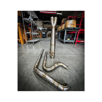 Metal pipes and a Y-shaped exhaust component rest on the concrete floor in a workshop environment, surrounded by shelves and boxes—a scene where SP Concepts works to perfect their Full Length Turnpike Works Edition for Harley '96-'16 Touring exhaust systems.