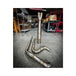 Metal pipes and a Y-shaped exhaust component rest on the concrete floor in a workshop environment, surrounded by shelves and boxes—a scene where SP Concepts works to perfect their Full Length Turnpike Works Edition for Harley &