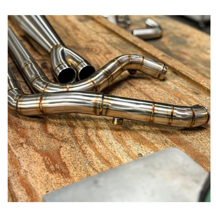 Metal pipes and a Y-shaped exhaust component rest on the concrete floor in a workshop environment, surrounded by shelves and boxes—a scene where SP Concepts works to perfect their Full Length Turnpike Works Edition for Harley &