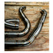 Metal pipes and a Y-shaped exhaust component rest on the concrete floor in a workshop environment, surrounded by shelves and boxes—a scene where SP Concepts works to perfect their Full Length Turnpike Works Edition for Harley &