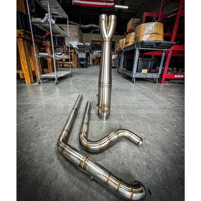 The SP Concepts Full Length Turnpike Works Edition for Harley '17-'24 M8 Touring headers and exhaust pipes made of stainless steel are neatly arranged on the warehouse floor, surrounded by shelves and boxes in the background.