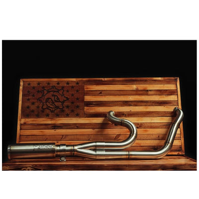 The Fab 28 Stainless Exhaust with Canister Muffler For Harley '18+ M8 Softail is showcased in front of a wooden panel that features a carved American flag and a design incorporating pipes and gears, echoing the craftsmanship found on a Harley M8 Softail.