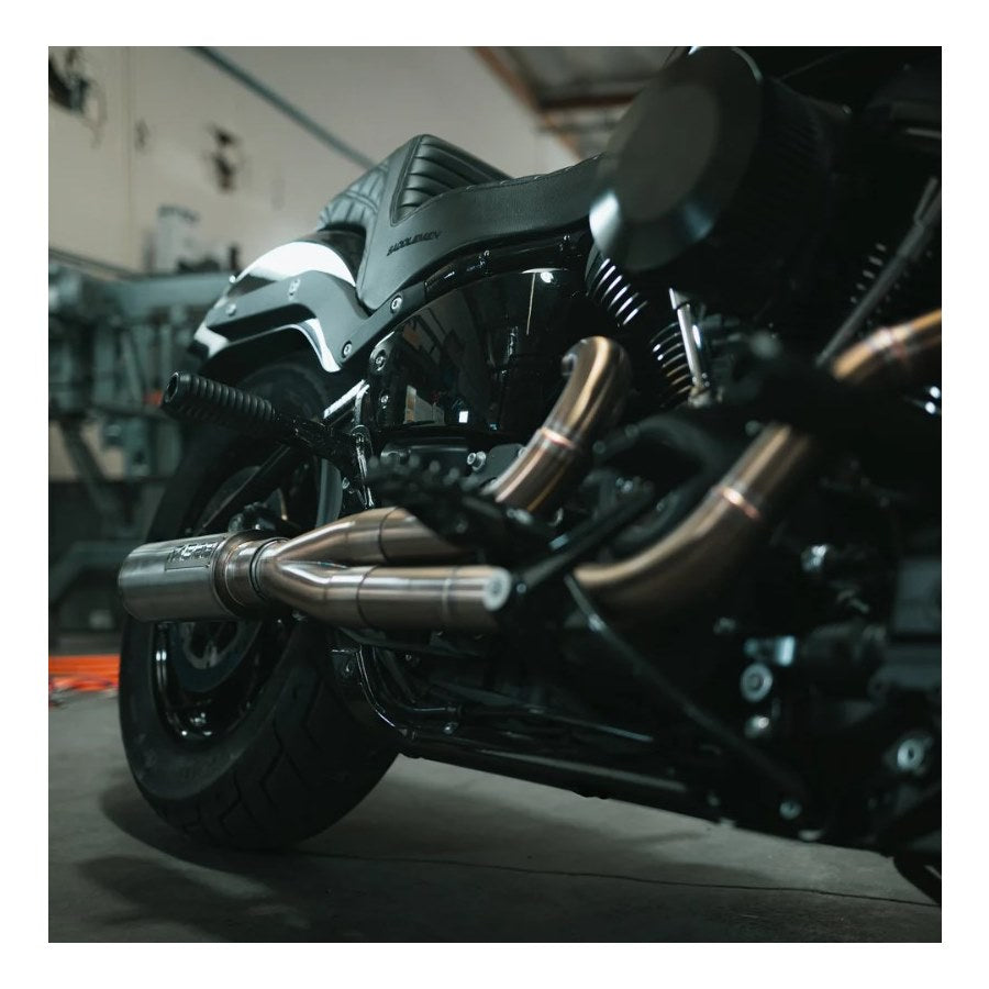 The Fab 28 Stainless Exhaust with Canister Muffler For Harley '18+ M8 Softail is showcased in front of a wooden panel that features a carved American flag and a design incorporating pipes and gears, echoing the craftsmanship found on a Harley M8 Softail.