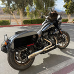 A sleek black Harley Davidson M8 Softail with an SP Concepts Works Edition Mid Length Elsinore Exhaust gleams under the sun on a concrete surface. Featuring saddlebags and a distinctive logo, this motorcycle exudes style and power.