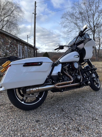 A dimly lit garage houses a gray motorcycle equipped with an SP Concepts Mid Length Elsinore Exhaust for Harley Davidson M8 Touring Bagger 2017-Up. Shelves surround it, with an American flag providing a striking backdrop.