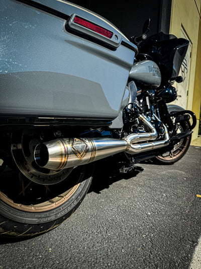 In a workshop with polished metal exhaust pipes, a worker, and an American flag above, the SP Concepts Works Edition Mid Length Elsinore Exhaust for Harley Davidson M8 Touring Bagger 2017-Up is showcased as ideal for enhancing performance.