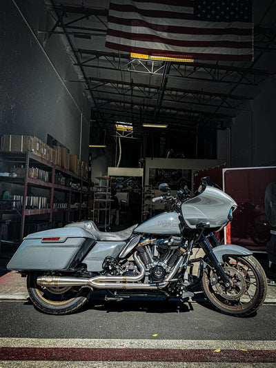 A dimly lit garage houses a gray motorcycle equipped with an SP Concepts Mid Length Elsinore Exhaust for Harley Davidson M8 Touring Bagger 2017-Up. Shelves surround it, with an American flag providing a striking backdrop.