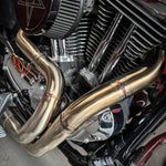 A SP Concepts stainless steel header with multiple bends rests on a gray surface, highlighting its craftsmanship. In the background, an American flag-designed welding mask nods to the customization spirit of Harley Davidson Sportster and Lanesplitter Exhaust fans.