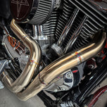 A SP Concepts stainless steel header with multiple bends rests on a gray surface, highlighting its craftsmanship. In the background, an American flag-designed welding mask nods to the customization spirit of Harley Davidson Sportster and Lanesplitter Exhaust fans.