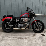 An SP Concepts Lanesplitter Exhaust for Harley Davidson Sportster (1986-2003) is showcased on a textured surface with intricate curves. Nearby, an American flag-patterned helmet hints at the increased torque this meticulously welded stainless steel product promises.