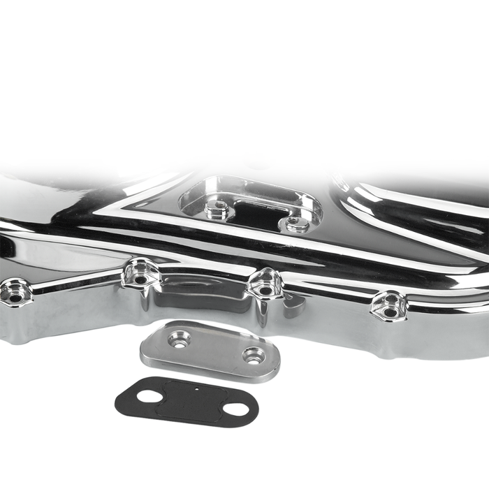 A close-up of a shiny metal engine or machine part, possibly from a Harley-Davidson Sportster, with two smaller metal and rubber gaskets—including a James Inspection Cover Gasket for 2004-2022 Harley-Davidson Sportster by James Gaskets—placed in front of it on a white background.