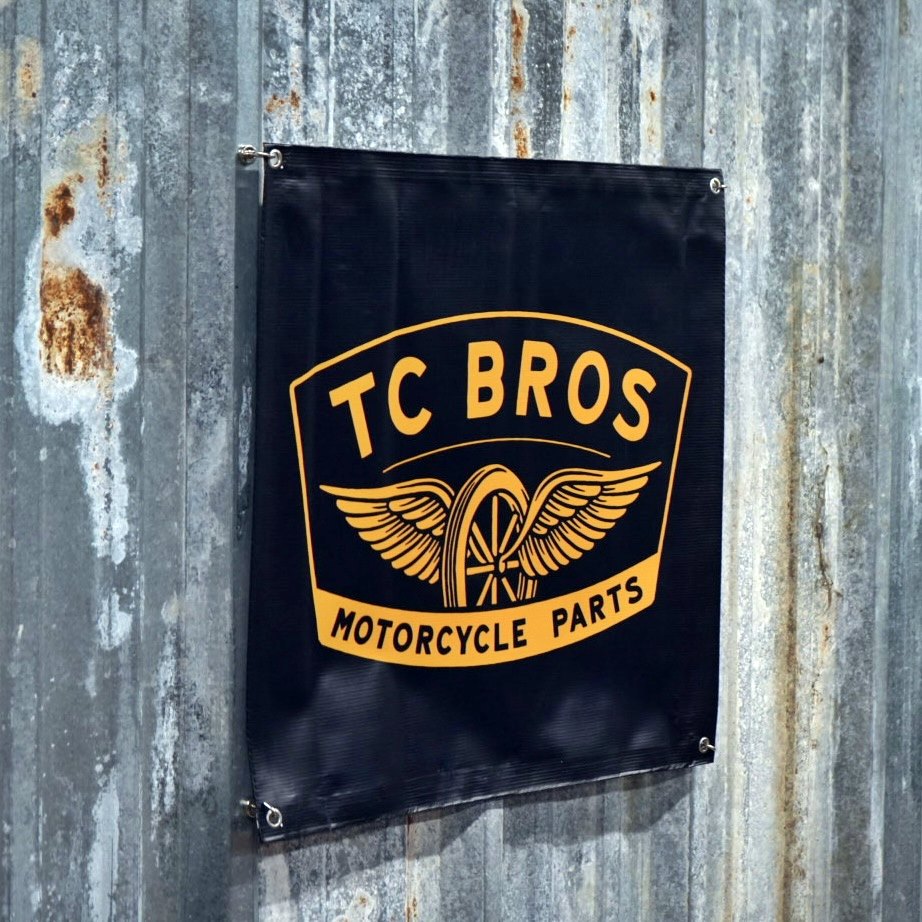 A TC Bros. Winged Wheel Banner 2ftx2ft featuring the logo "TC Bros Motorcycle Parts" with winged graphics, mounted on a weathered gray metal wall, equipped with metal grommets.