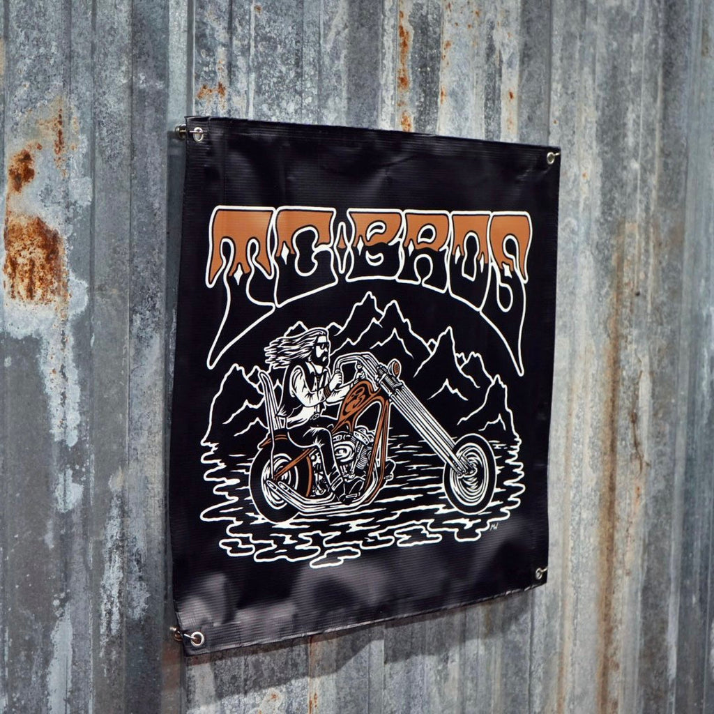 A black, weatherproof flag with a TC Bros. logo and a graphic of a skeleton on a motorcycle, mounted on a rusted metal wall.
