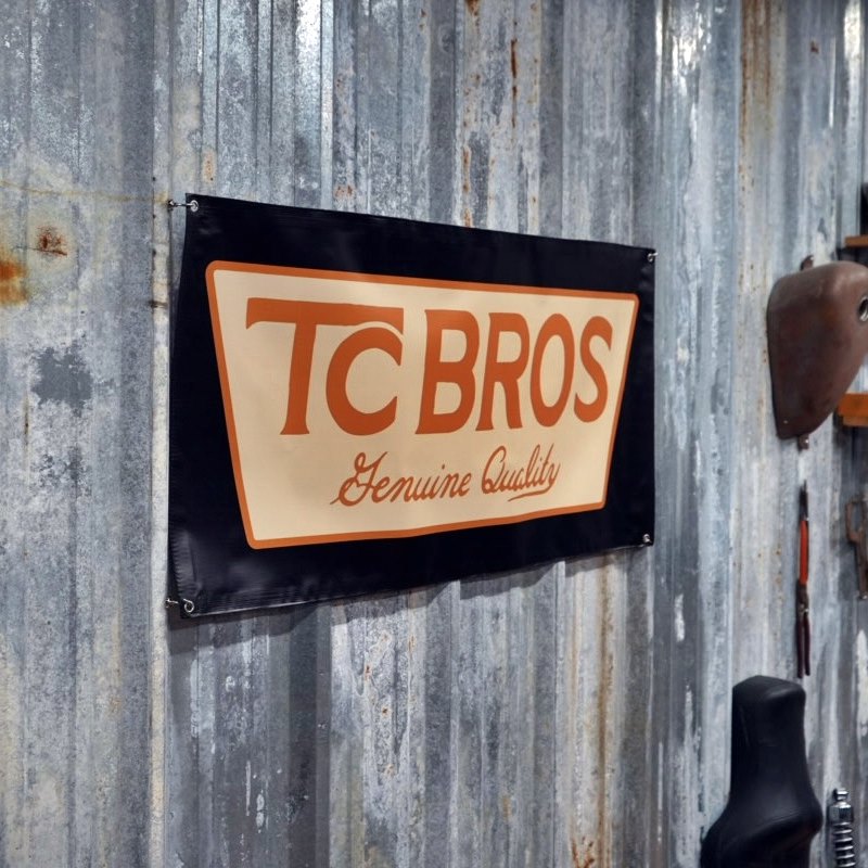 A TC Bros. Genuine Quality Banner 2ftx4ft from TC Bros. on a corrugated metal wall in a workshop environment.