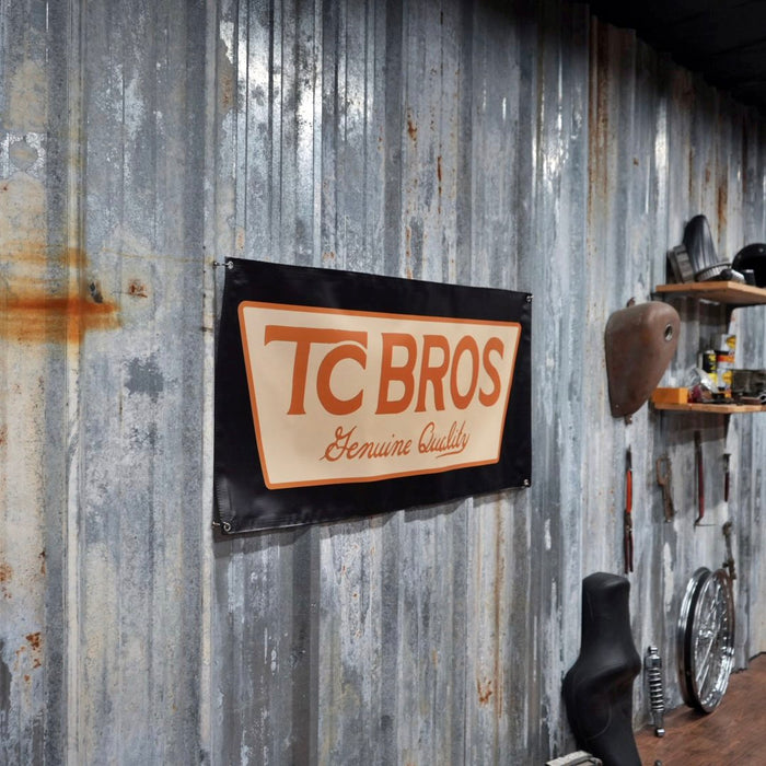 A TC Bros. Genuine Quality Banner 2ftx4ft from TC Bros. on a corrugated metal wall in a workshop environment.