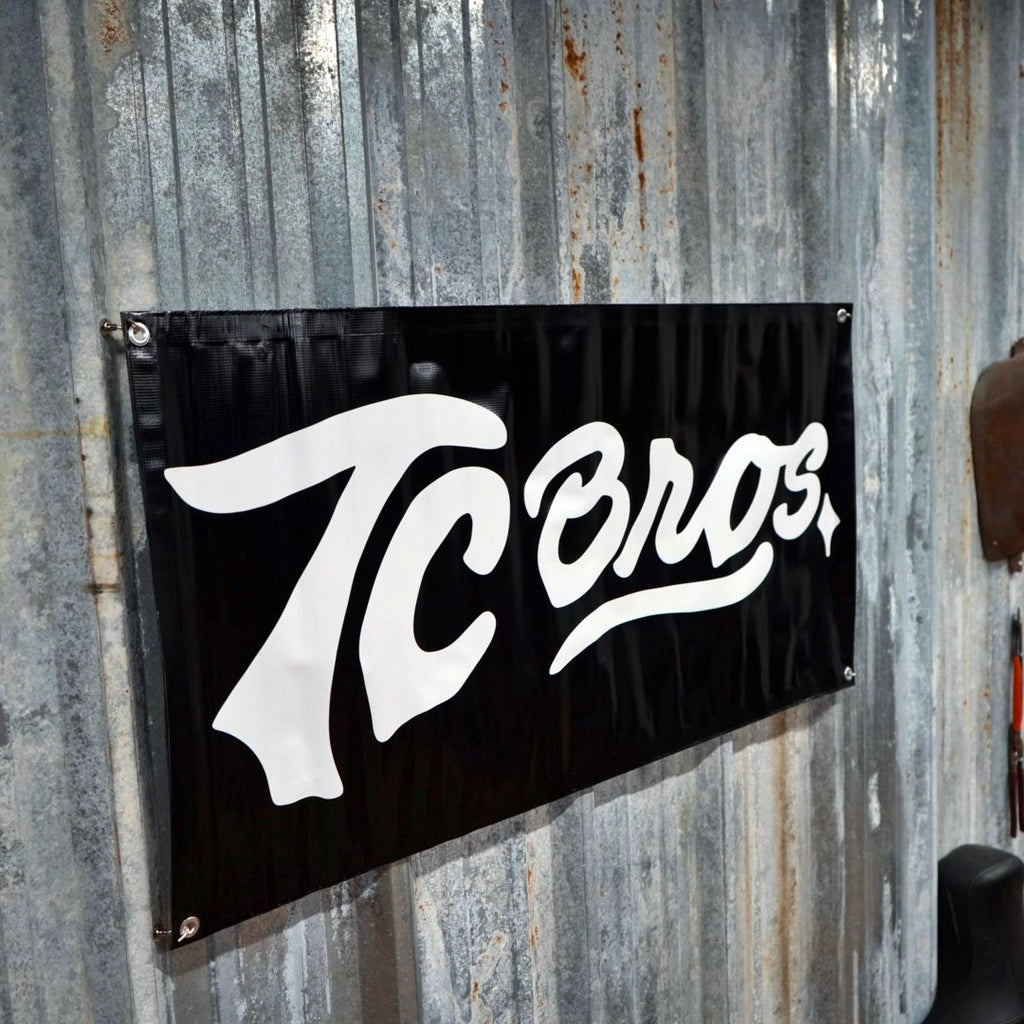 Black TC Bros. Banner 2ft x 4ft with the word "tcoros" in white cursive font, mounted on a corrugated metal wall.