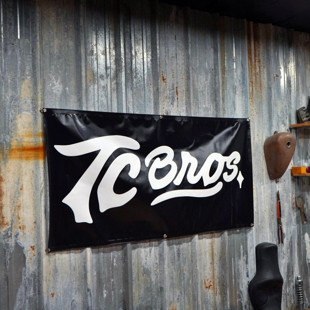 A black TC Bros. vinyl banner with "tc bros" in white script, mounted on a corrugated metal wall in a garage.